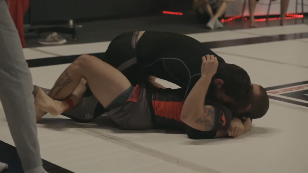 two fighters lying on the floor and hugging, one in black sport suit, another in gray and red