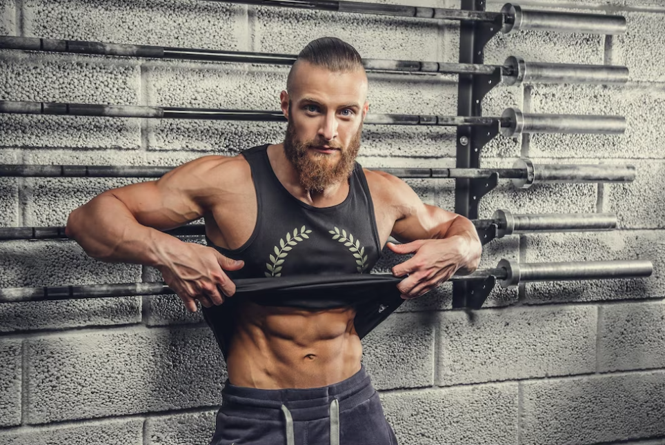 bearded muscular man lifts his gray t-shirt showing his abs in front of the wall